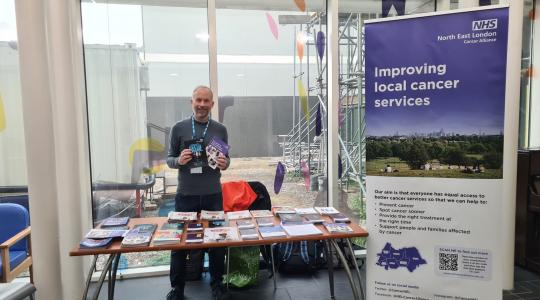 A man is standing behind a table which has cancer information leaflets on it. On the right is a banner which says Improving Local Cancer Services