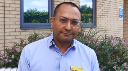 The image is of a male, asian doctor standing outside a brick building with windows