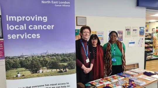 Three ladies are standing behind a table which has cancer information leaflets on it. A pop up banner says 'Improving Local Cancer Services' on it.