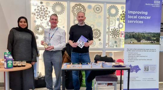 Two men and a lady are standing in front of a cancer awareness information stand. Cancer leaflets are displayed on a table. A pop-up banner says 'Improving local cancer services' on it.