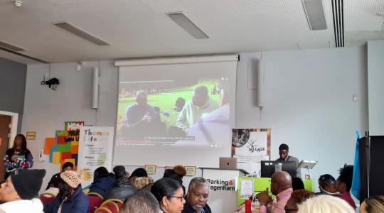 A video on prostate cancer is being shown on a big screen in front of an audience in a large room