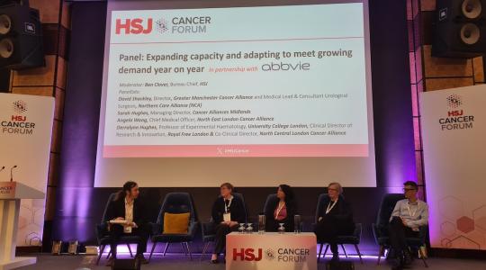 5 people are sat on a stage as a panel of speakers with a large screen behind them which says HSJ Cancer Forum on it