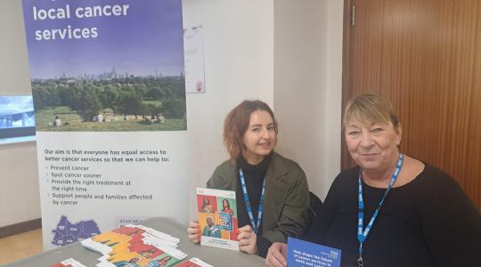 Two women are sat at a table which has cancer information leaflets on it. To the left of them is a pop up banner which says 'Improving local cancer services' on it