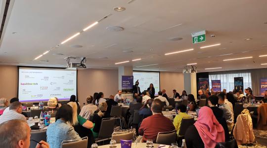 The image is of a full conference theatre with a mix of adults sitting around tables facing a screen and presenter.