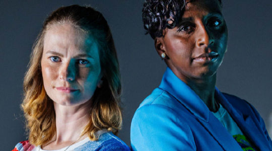 Two female athletes are standing next to each other. One is white and has long brown hair. The other is black and has short black hair.