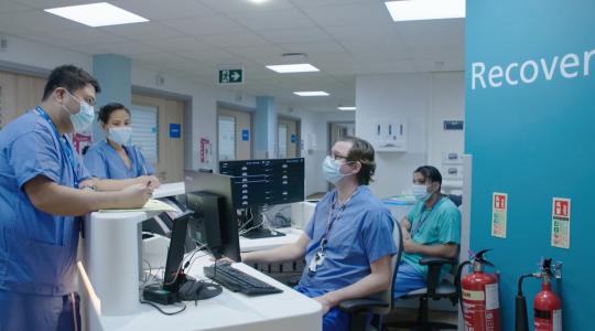 The image is of a reception in a hospital and features four staff wearing surgical gowns and masks