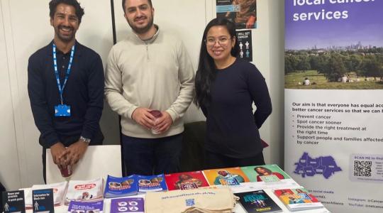 Three people are standing behind a table which has a range of cancer information leaflets on it.