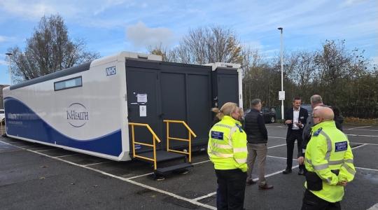 The image is of a lung scanning truck in a supermarket car park. Standing outside are a group of people talking