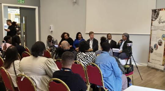 A panel of speakers is sat down in front of an audience, who are also seated, in a large meeting room.