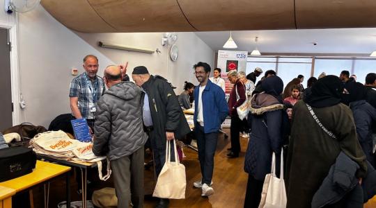 A large number of people is shown in a community hall. There is a group of people chatting to a man behind a table with information leaflets on it.