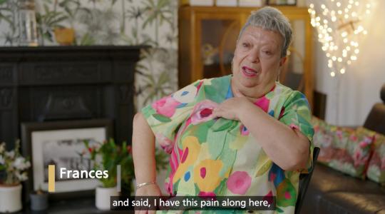 A lady in a flowery dress is sitting in her home and pointing to her shoulder. A caption below says "and said, I have this pain along here"