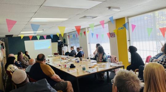 A large meeting room has a long table in the centre and people are sat around the table looking at the presentation screen, which is at the far end
