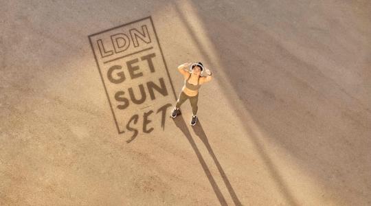 A runner is standing on a sandy background giving the thumbs up to the sky. On the sand is written LDN Get Sun Set.