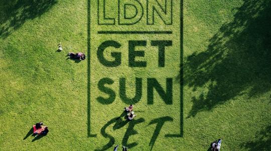 An overhead shot of a sunny park with people sitting on the grass. On the grass are the words LDN Get Sun Set.