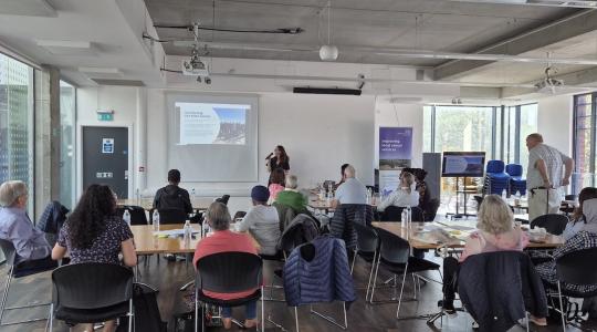 A lady is presenting to an audience who are sat around tables in a large room. She is holding a microphone and standing by a presentation screen