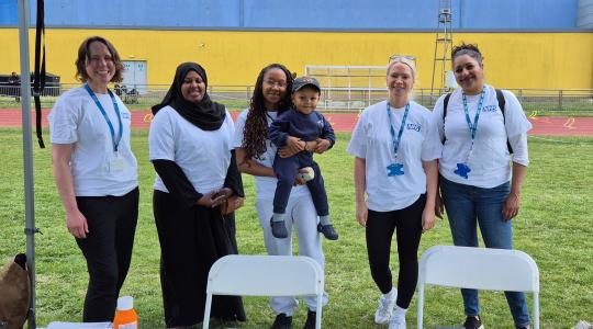 Five females wearing white T-shirts are standing outside under a gazebo behind a table which has cancer information leaflets on it. The lady in the middle is holding a small child.