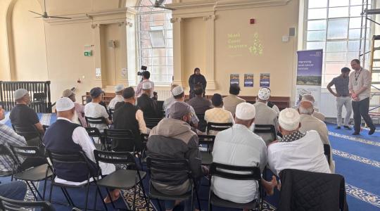 An audience is sat watching a presentation taking place in a Mosque.