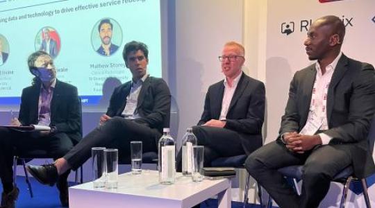 Four men in suits are sat on a stage as part of a speaker panel at an event, with a large presentation screen behind them.