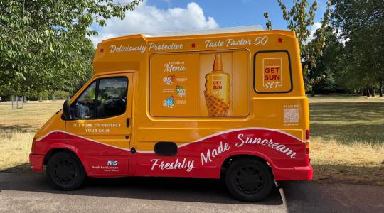 An ice cream van is stationary in a park and has been repainted in bright yellow. Instead of advertising ice cream it is promoting sun cream.