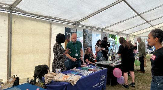 Two people are standing behind a table which has cancer information leaflets on it. They are inside a large tent in a park.