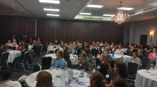 A large hall is filled with round tables and guests sitting at them for a conference.