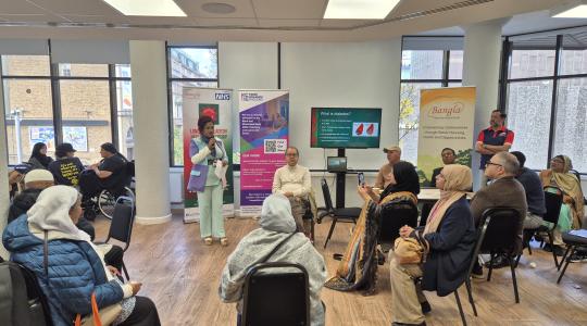 A woman is standing in the middle of a room giving a presentation to a number of people who are sat down in chair facing her.