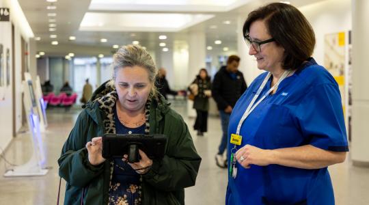 A nurse is standing next to a lady who is looking at the screen on a handheld digital device.