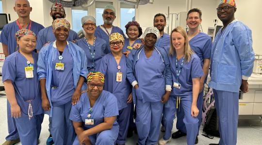 A team of clinicians in hospital scrubs is standing in a hospital operating theatre for a team photo.