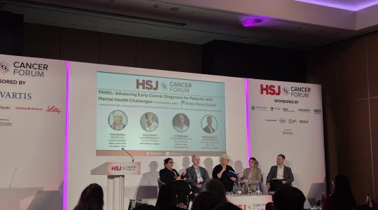 A panel of speakers are sat on a stage with a presentation screen behind them displaying the words HSJ Cancer Forum.