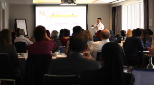A man is standing at the back of a room doing a presentation in front of a large audience who are seated around circular tables.