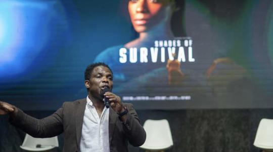 A man is standing in front of a title of a movie which says Shades of Survival.
