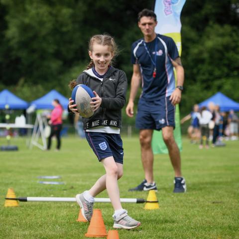 A young girl is holding a rugby ball and a man is standing in the background coaching her.