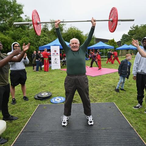 An old man is lifting some weights in a park with people watching and cheering him on.