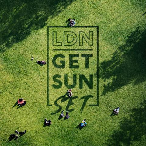 An overhead shot of a sunny park with people sitting on the grass. On the grass are the words LDN Get Sun Set.
