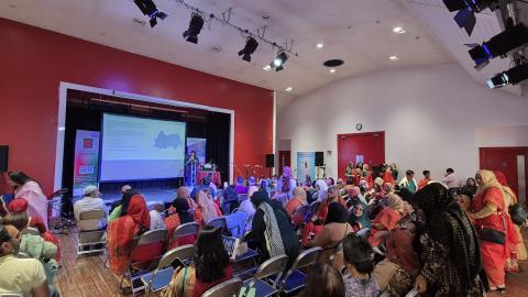 A lady is on stage delivering a presentation to a large audience who are seated in a community hall.