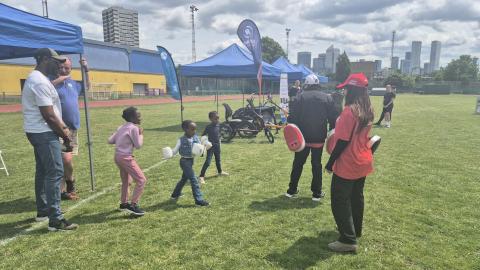 A couple of young children are being given boxing lessons by some adults in a field on a sunny day.