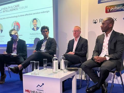 Four men in suits are sat on a stage as part of a speaker panel at an event, with a large presentation screen behind them.