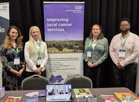 Four people are standing next to a banner which says improving local cancer services on it. They are standing behind a table which has cancer information leaflets on it.