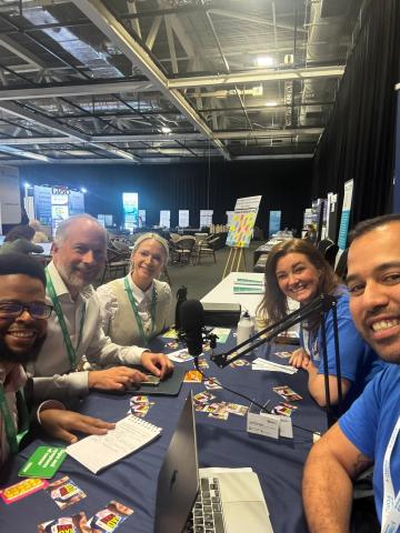 Five people are sat at a table in a large exhibition hall recording a podcast.