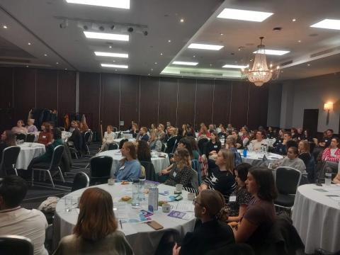 A large hall is filled with round tables and guests sitting at them for a conference.