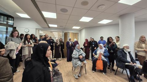 A large number of people are sat down and standing in a room facing to the front of the room.