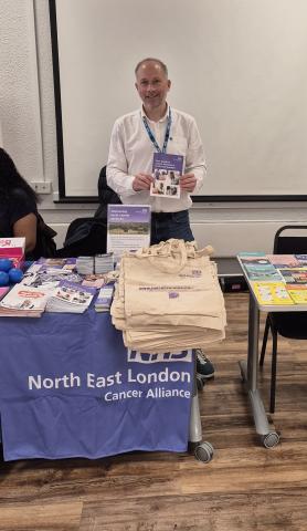 A man is standing behind a table with a lot of leaflets and information on it and he is holding up one of the leaflets.