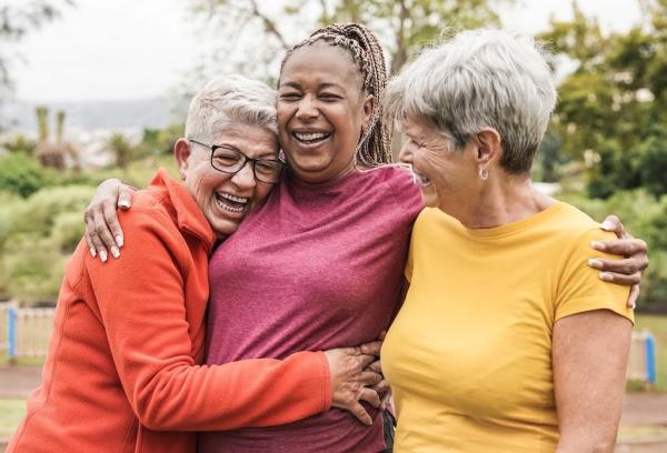 Three ladies wearing bright coloured tops are hugging each other outside in a park
