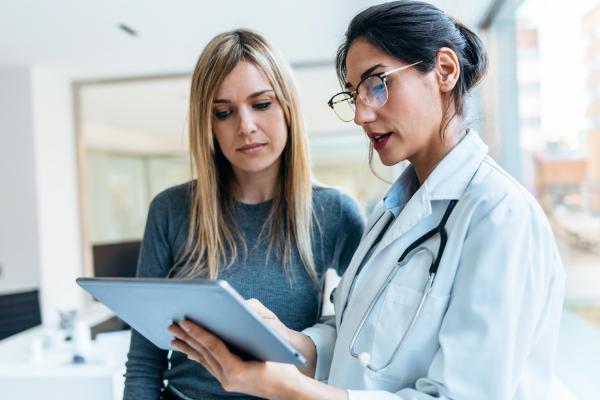 A lady with long light brown hair is talking to a healthcare professional holding a tablet and wearing glasses and a white jacket.