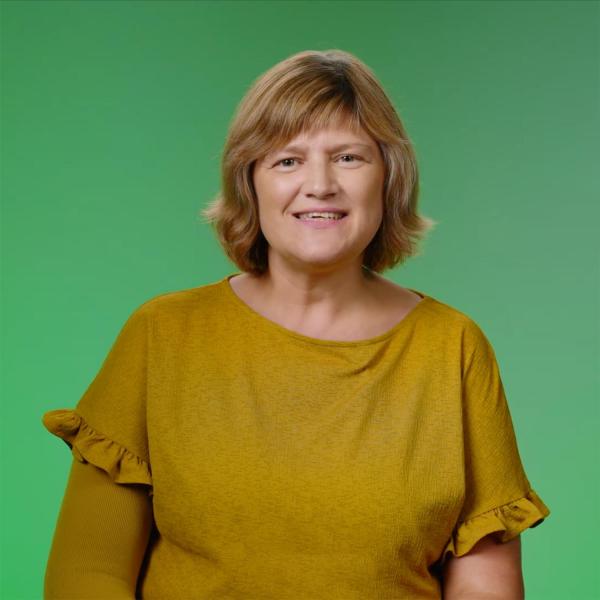 A lady with light brown hair and wearing a mustard colour top is standing in front of a bright green background