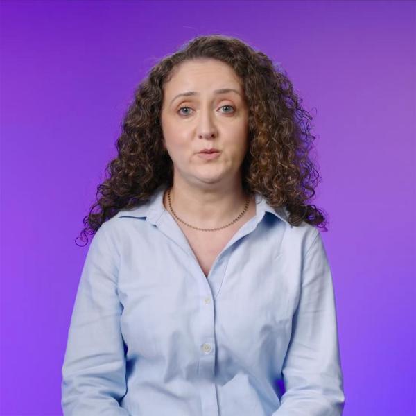 A lady with brown curly hair wearing a pale shirt is standing in front of a bright purple background