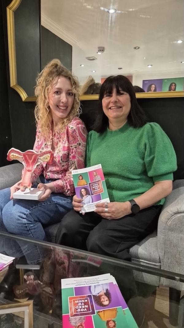 Two women are sat on a sofa. One is holding a leaflet and the other a model of ovaries.