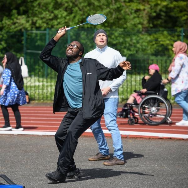 A man is playing badminton and behind him is a another man, as well as a lady in a wheelchair.