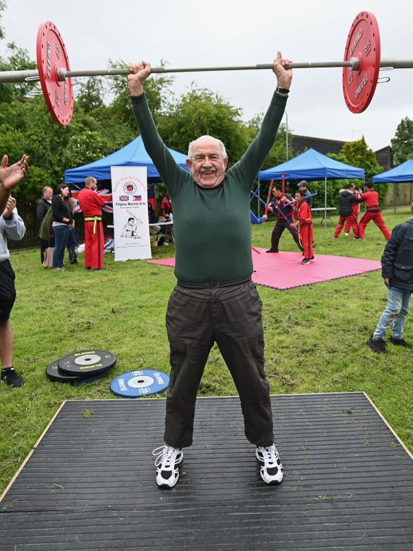 An old man is lifting a set of weights above his head in a park.