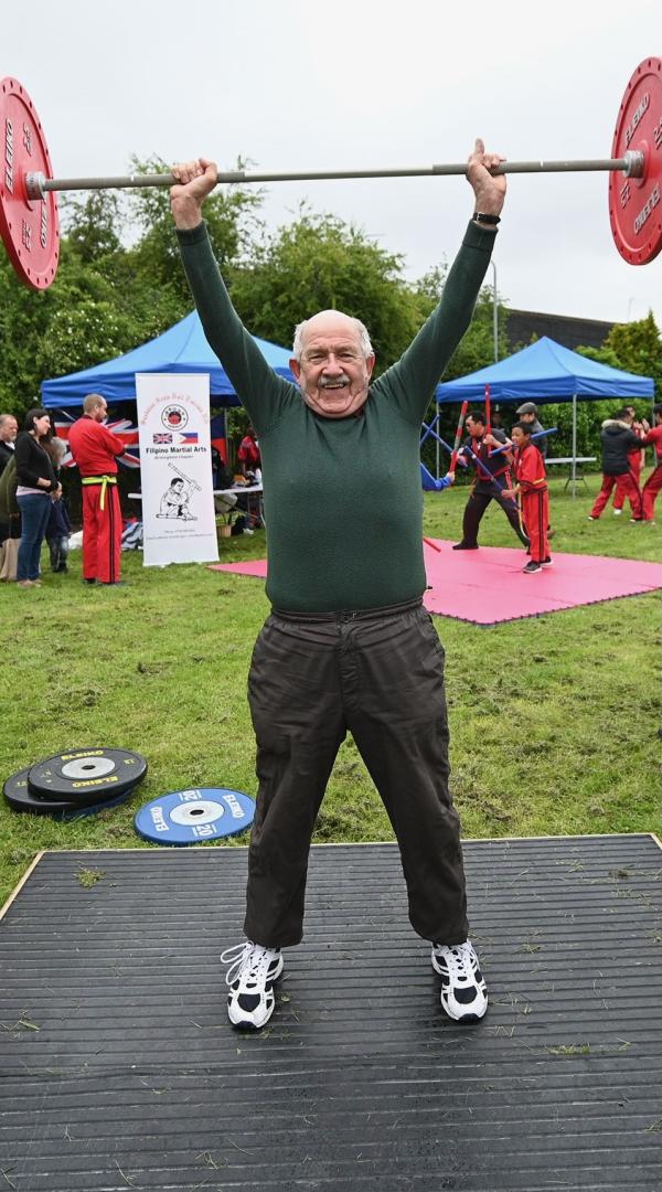 An old man is lifting a set a weights above his head in a park.